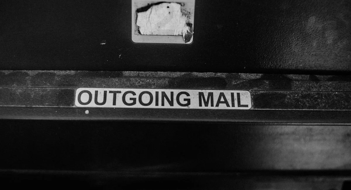 Black and white photograph of an 'Outgoing Mail' slot with rustic textures.